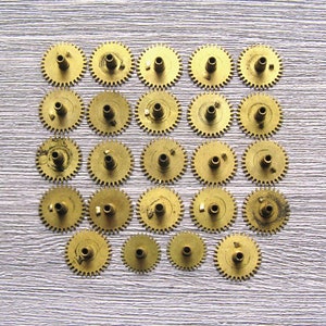 May include: A collection of 25 gold-colored metal gears arranged in a square pattern on a light gray textured surface.