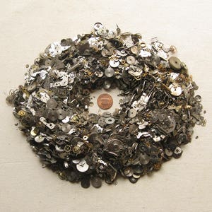 Op de afbeelding: Een cirkelvormige opstelling van kleine, metalen horlogeonderdelen, tandwielen en componenten. De stukken zijn voornamelijk zilver en goud van kleur, met een cent in het midden. De componenten zijn gerangschikt in een kransachtige vorm.