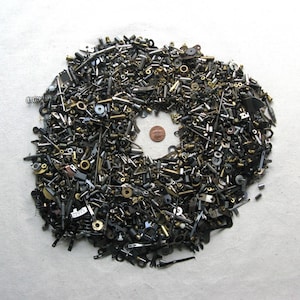 Puede incluir: Un montón grande de tornillos, tuercas, pernos y otras piezas de ferretería de metal en tonos plateados y dorados. Una sola moneda de cobre está en el centro del montón.