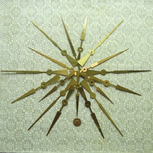 May include: A set of 18 gold-coloured clock hands of various sizes, arranged in a starburst pattern on a white fabric background.