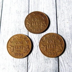 Może przedstawiać: Trzy brązowe kanadyjskie monety jednocentowe. Każda moneta zawiera napis "CANADA ONE CENT" oraz rok wybicia, 1928 lub 1932, wraz z motywami liści klonu. Monety ułożone są na jasnej drewnianej powierzchni.