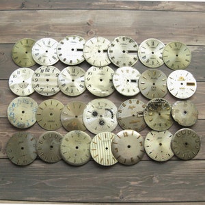 May include: A collection of 25 vintage watch faces, most with Roman numeral markings, in various styles and colors. The watch faces are arranged on a wooden surface.