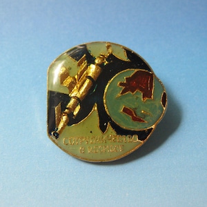 Puede incluir: Un pin redondo de color dorado con un diseño azul, verde y rojo. El pin presenta un cohete y un globo terráqueo. El texto "СОТРУДНИЧЕСТВО В КОСМОСЕ" está escrito en letras doradas alrededor de la parte inferior del pin.