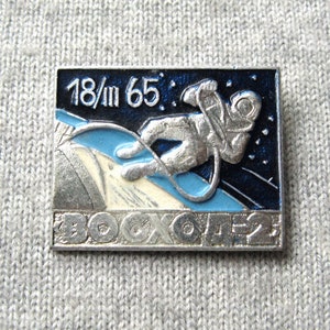 Puede incluir: Un pin de esmalte plateado y azul con un astronauta en un traje espacial flotando en el espacio. El pin tiene el texto "18/m 65" y "BOCCOTEZ".