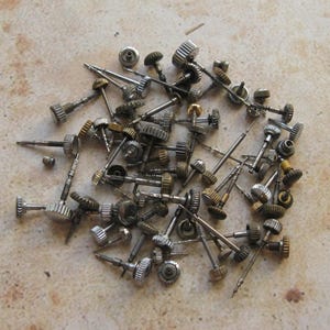 Op de afbeelding: Een verzameling vintage horlogeonderdelen, waaronder tandwielen, stengels en kronen. De onderdelen zijn van metaal en hebben een zilveren en gouden afwerking.