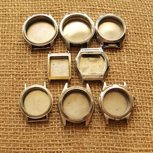 May include: A collection of seven empty watch cases in various shapes, including round, square, and rectangular. The cases are made of silver-colored metal and are arranged on a textured, tan surface.
