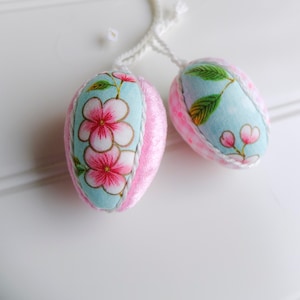 Könnte beinhalten: Zwei rosa und blaue Stoff-Ostereier mit Blumenmustern. Die Eier hängen an einer weißen Schnur.