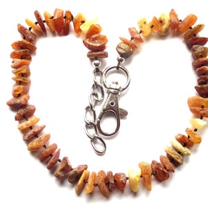 May include: A necklace made of amber stones in shades of brown, orange, and yellow, strung together with a silver-colored clasp. The necklace is arranged in a heart shape against a white background.