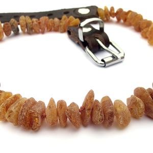 May include: A necklace featuring raw amber stones in shades of brown and orange. The amber beads are strung together, with a dark brown leather strap and a silver-colored metal clasp.