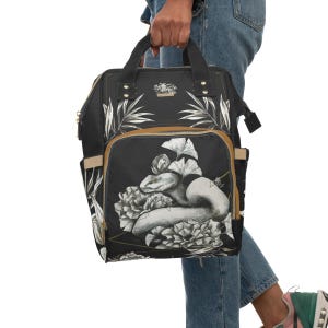 Puede incluir: Bolso de pañales negro con una serpiente blanca y un diseño floral. El bolso tiene una cremallera marrón y un acento dorado.