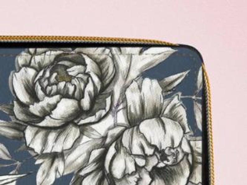 K&ouml;nnte beinhalten: Nahaufnahme einer dunkelblauen Geldb&ouml;rse mit goldenem Rei&szlig;verschluss und floralem Design. Das Design zeigt gro&szlig;e, detaillierte wei&szlig;e und graue Pfingstrosenbl&uuml;ten und -bl&auml;tter. Die Geldb&ouml;rse befindet sich auf einem rosa Hintergrund.