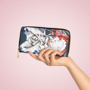 Puede incluir: Una cartera azul oscuro con cremallera y una ilustración en acuarela. La obra de arte representa personajes de una película de animación, con una cremallera dorada y un fondo rosa. La cartera se sostiene en una mano.