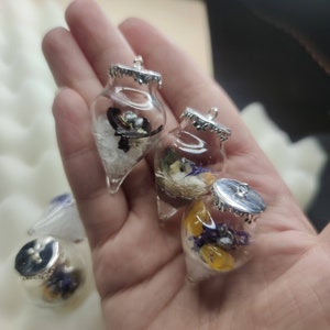 Puede incluir: Tres adornos de vidrio con tapas plateadas, cada uno lleno de flores secas y cristales. Los adornos tienen forma de lágrima y tienen un aspecto delicado y caprichoso.