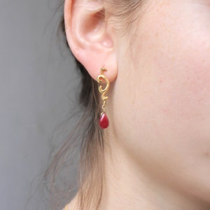 May include: Gold earrings with a dangling red gemstone. The earrings feature a delicate, swirling design.