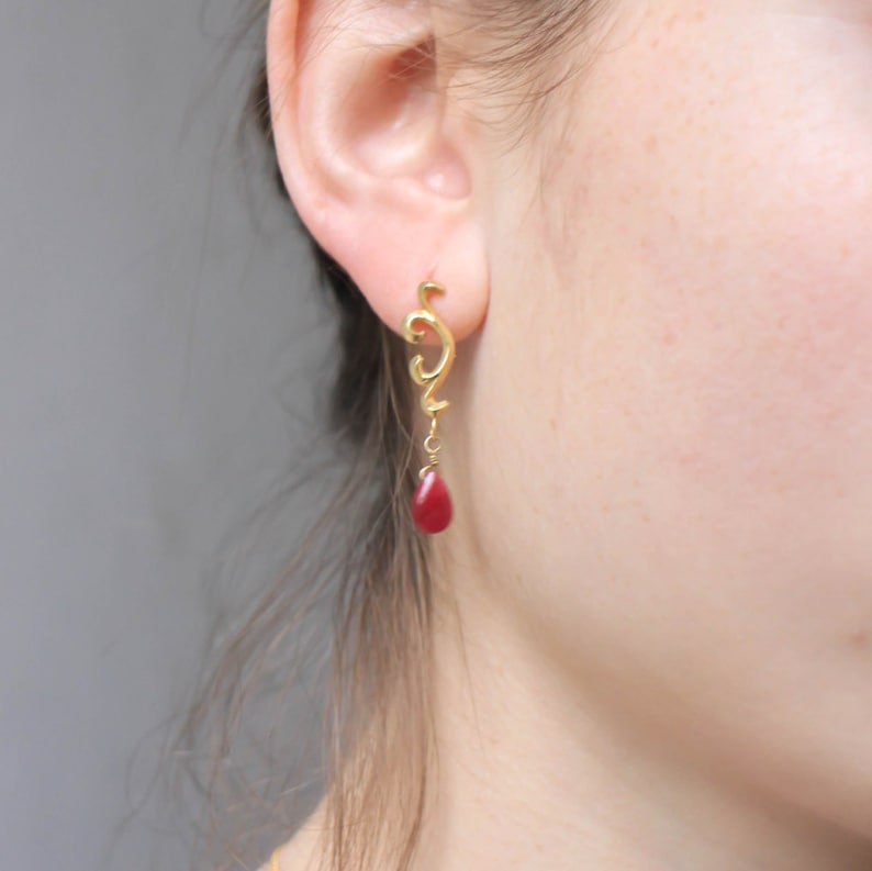 May include: Gold earrings with a dangling red gemstone. The earrings feature a delicate, swirling design.