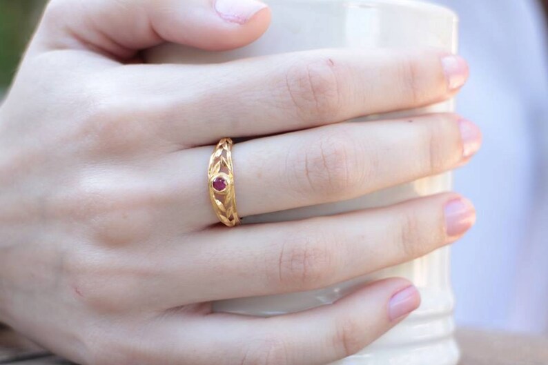 May include: A gold ring with a delicate, intricate design featuring a single red gemstone in the center.