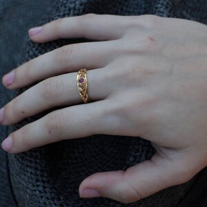 May include: A gold ring with a red gemstone set in the center. The ring has a braided design.