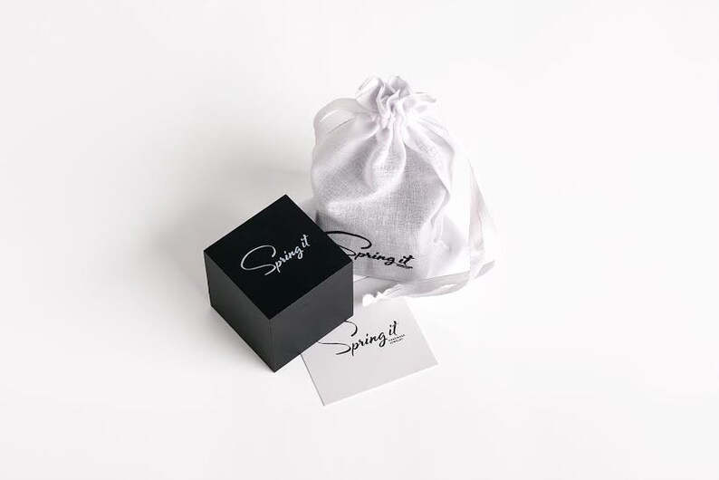 May include: A black square box with the text "Spring it" in white lettering. The box is sitting on a white surface next to a white drawstring bag with the text "Spring it" in black lettering. There is also a white card with the text "Spring it" in black lettering.