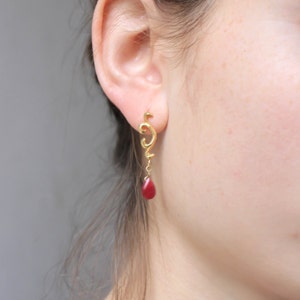 May include: Gold dangle earrings with a red teardrop-shaped stone. The earrings feature a delicate, swirling design.