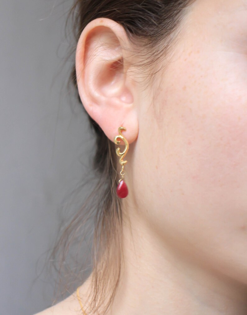 May include: Gold dangle earrings with a red teardrop-shaped stone. The earrings feature a delicate, swirling design.