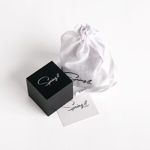 May include: A black square box with the text "Spring it" in white lettering. The box is sitting on a white surface next to a white drawstring bag with the text "Spring it" in black lettering. A white card with the text "Spring it" in black lettering is in front of the box.