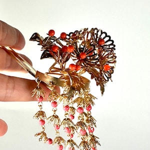 Op de afbeelding: Gouden haar kam met een bloemmotief en koraalkleurige kralen. De kam heeft een lange, hangende ketting van kralen en bladeren.