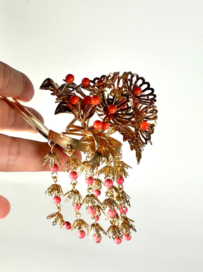 Op de afbeelding: Gouden haar kam met een bloemmotief en koraalkleurige kralen. De kam heeft een lange, hangende ketting van kralen en bladeren.