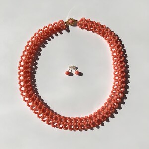 Puede incluir: Un collar de cuentas de color coral y un juego de pendientes a juego. El collar es un dise&ntilde;o de m&uacute;ltiples hilos con un cierre dorado. Los pendientes son peque&ntilde;os, redondos y de color coral. El conjunto de joyas se muestra sobre una superficie blanca.