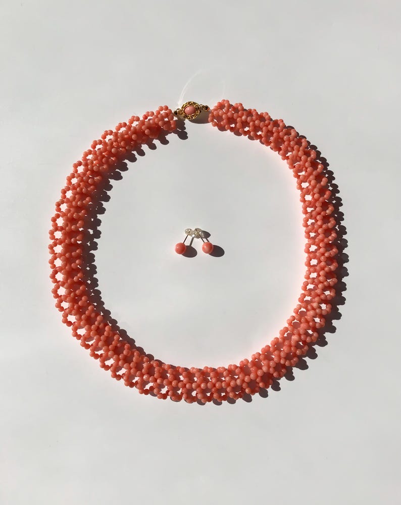 Puede incluir: Un collar de cuentas de color coral y un juego de pendientes a juego. El collar es un dise&ntilde;o de m&uacute;ltiples hilos con un cierre dorado. Los pendientes son peque&ntilde;os, redondos y de color coral. El conjunto de joyas se muestra sobre una superficie blanca.