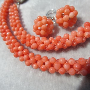 Op de afbeelding: Een sieradenset bestaande uit een ketting en een paar oorbellen, beide gemaakt van kleine, ronde, zalmroze kralen gerangschikt in een dicht, gestructureerd, bolvormig en touwachtig patroon, voorzien van zilverkleurige metalen onderdelen.