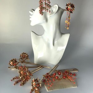 Op de afbeelding: Set van zes gouden haar kammen met koraal gekleurde kralen en bloemmotieven. De kammen zijn gerangschikt op een wit oppervlak.