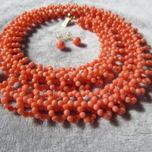 Puede incluir: Un collar de cuentas de color coral y un conjunto de pendientes a juego. El collar es un dise&ntilde;o de m&uacute;ltiples hilos con un cierre dorado. Los pendientes son peque&ntilde;as cuentas redondas. El conjunto de joyas se muestra sobre una superficie gris claro.