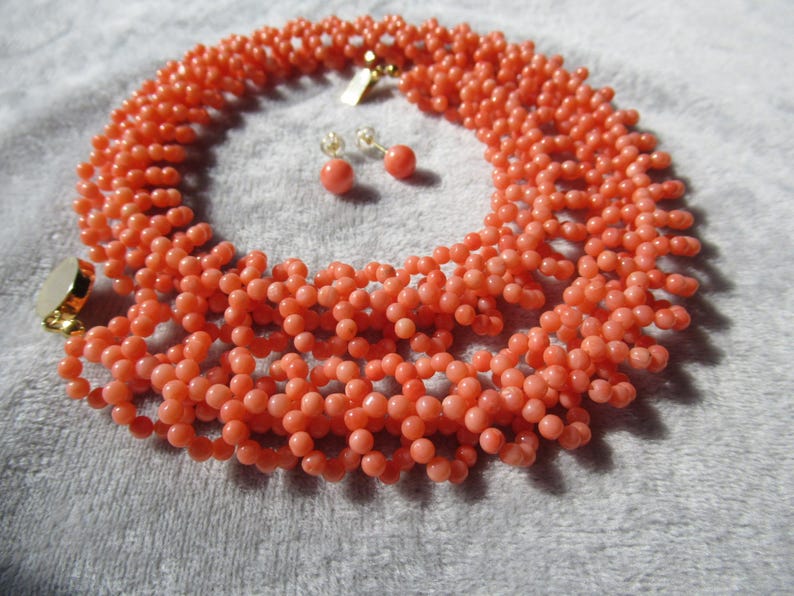 Puede incluir: Un collar de cuentas de color coral y un conjunto de pendientes a juego. El collar es un dise&ntilde;o de m&uacute;ltiples hilos con un cierre dorado. Los pendientes son peque&ntilde;as cuentas redondas. El conjunto de joyas se muestra sobre una superficie gris claro.