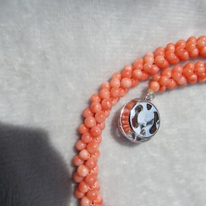 Op de afbeelding: Een ketting bestaande uit meerdere strengen kleine, ronde, zalmroze kralen geweven in een gedraaid touwontwerp. De ketting heeft een ronde, zilverkleurige metalen sluiting met een zichtbare stempel met de tekst SILVER.