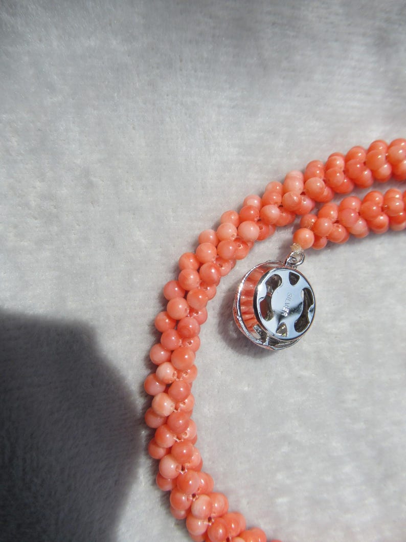 Op de afbeelding: Een ketting bestaande uit meerdere strengen kleine, ronde, zalmroze kralen geweven in een gedraaid touwontwerp. De ketting heeft een ronde, zilverkleurige metalen sluiting met een zichtbare stempel met de tekst SILVER.