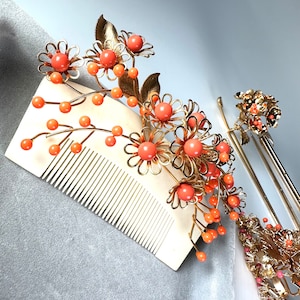 Op de afbeelding: Twee gouden haar kammen met oranje bloem accenten. De kammen zijn versierd met kleine, ronde oranje kralen en gouden bladeren. De kammen liggen op een grijze achtergrond.