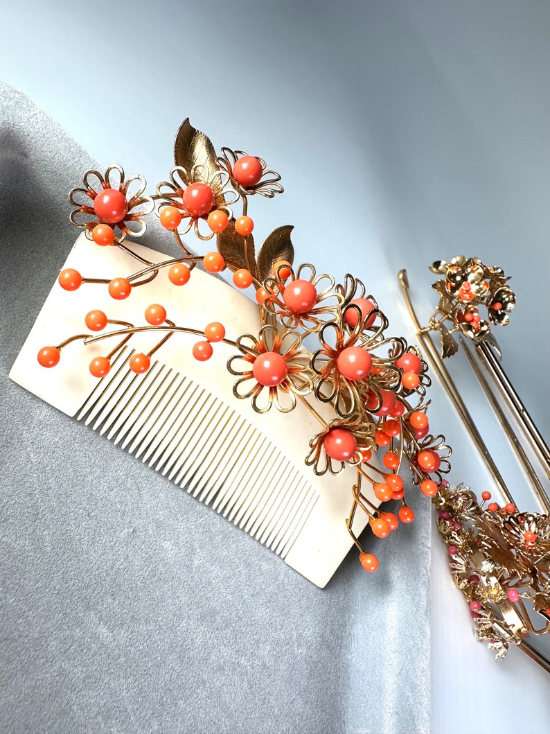 Op de afbeelding: Twee gouden haar kammen met oranje bloem accenten. De kammen zijn versierd met kleine, ronde oranje kralen en gouden bladeren. De kammen liggen op een grijze achtergrond.