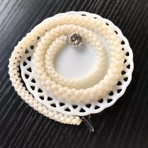May include: A white pearl necklace with a silver clasp. The necklace is layered and displayed on a white plate with a scalloped edge.