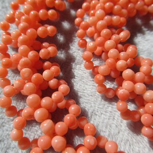 Puede incluir: Primer plano de un collar de coral con hebras de peque&ntilde;as cuentas redondas de color melocot&oacute;n. Las cuentas est&aacute;n ensartadas, creando una apariencia texturizada y org&aacute;nica. El collar se muestra sobre un fondo suave y gris claro.