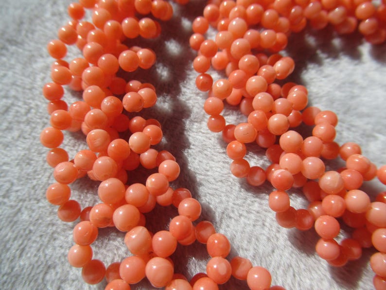 Puede incluir: Primer plano de un collar de coral con hebras de peque&ntilde;as cuentas redondas de color melocot&oacute;n. Las cuentas est&aacute;n ensartadas, creando una apariencia texturizada y org&aacute;nica. El collar se muestra sobre un fondo suave y gris claro.
