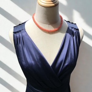 Op de afbeelding: Een mouwloze marineblauwe jurk met een V-hals en geplooide schouderdetails, gepresenteerd op een paspop. De jurk is gecombineerd met een korte, ronde ketting gemaakt van kleine, getextureerde, koraalkleurige kralen die nauw aansluiten.