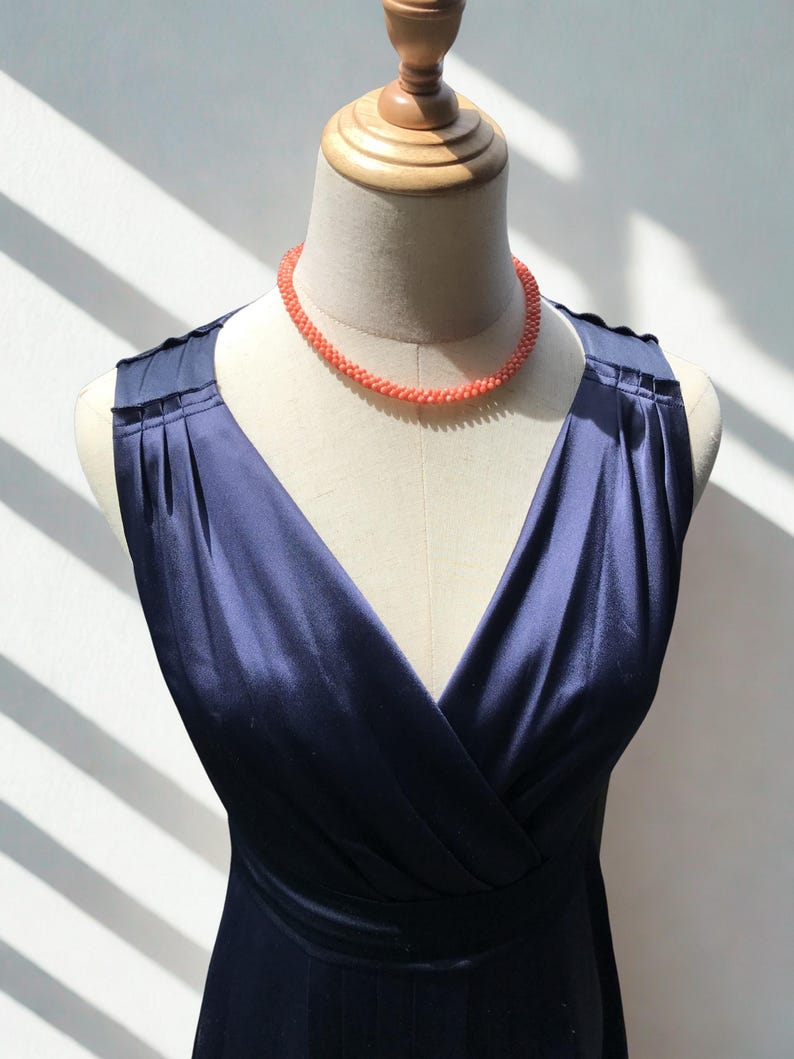 Op de afbeelding: Een mouwloze marineblauwe jurk met een V-hals en geplooide schouderdetails, gepresenteerd op een paspop. De jurk is gecombineerd met een korte, ronde ketting gemaakt van kleine, getextureerde, koraalkleurige kralen die nauw aansluiten.