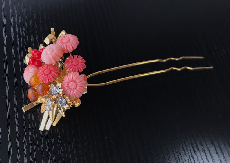 Puede incluir: Una horquilla de pelo dorada con un grupo de flores rosas y rojas, cuentas de &aacute;mbar y diamantes de imitaci&oacute;n.