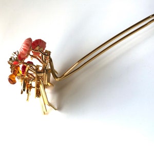 Puede incluir: Una horquilla de pelo dorada con un dise&ntilde;o floral decorativo que presenta detalles en rosa, naranja y blanco. La horquilla est&aacute; hecha de metal y tiene un dise&ntilde;o simple y elegante.