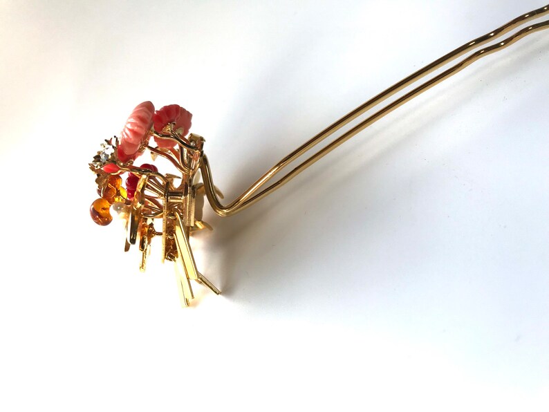 Puede incluir: Una horquilla de pelo dorada con un dise&ntilde;o floral decorativo que presenta detalles en rosa, naranja y blanco. La horquilla est&aacute; hecha de metal y tiene un dise&ntilde;o simple y elegante.