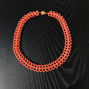 Puede incluir: Un collar de cuentas de color coral con un cierre dorado. El collar est&aacute; compuesto por peque&ntilde;as cuentas redondas dispuestas en un patr&oacute;n geom&eacute;trico. El cierre presenta una peque&ntilde;a piedra redonda de color coral. El collar se muestra sobre una superficie oscura.