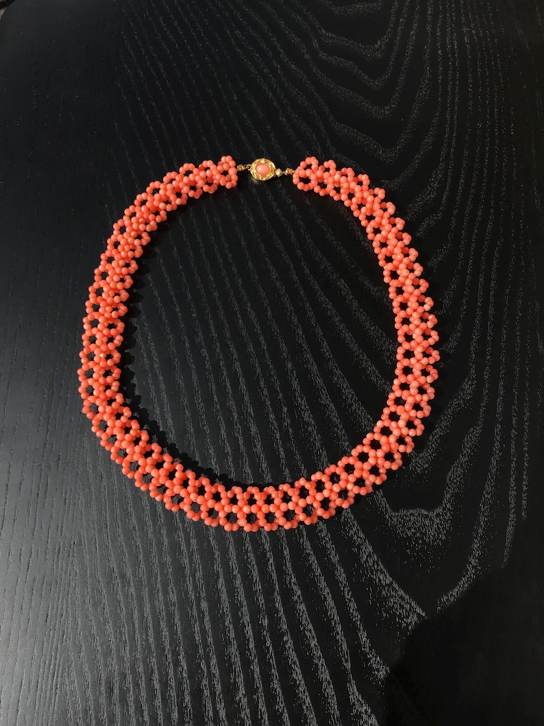 Puede incluir: Un collar de cuentas de color coral con un cierre dorado. El collar est&aacute; compuesto por peque&ntilde;as cuentas redondas dispuestas en un patr&oacute;n geom&eacute;trico. El cierre presenta una peque&ntilde;a piedra redonda de color coral. El collar se muestra sobre una superficie oscura.
