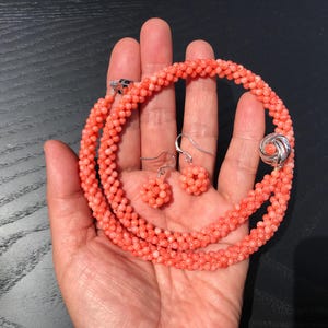 Op de afbeelding: Een zalmkleurige kralenketting en een bijpassend paar oorbellen. De ketting is gemaakt van kleine, getextureerde bolvormige kralen die in een doorlopende streng zijn gerangschikt, vastgezet met een zilverkleurige ronde sluiting met een centrale kraal.