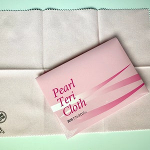 以下が含まれることがあります： 「Pearl Teri Cloth」と日本語で「真珠でクロス」と書かれたピンクのパッケージ。パッケージは、「Pearl Science Laboratory」の文字とロゴが入った白い布の上に置かれている。