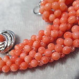 Op de afbeelding: Een meerstrengs ketting bestaande uit kleine, ronde, zalmkleurige kralen. De ketting heeft een ronde, zilverkleurige metalen sluiting met een gestructureerd, wervelend ontwerp en een bijpassende zalmkleurige kraal in het midden.